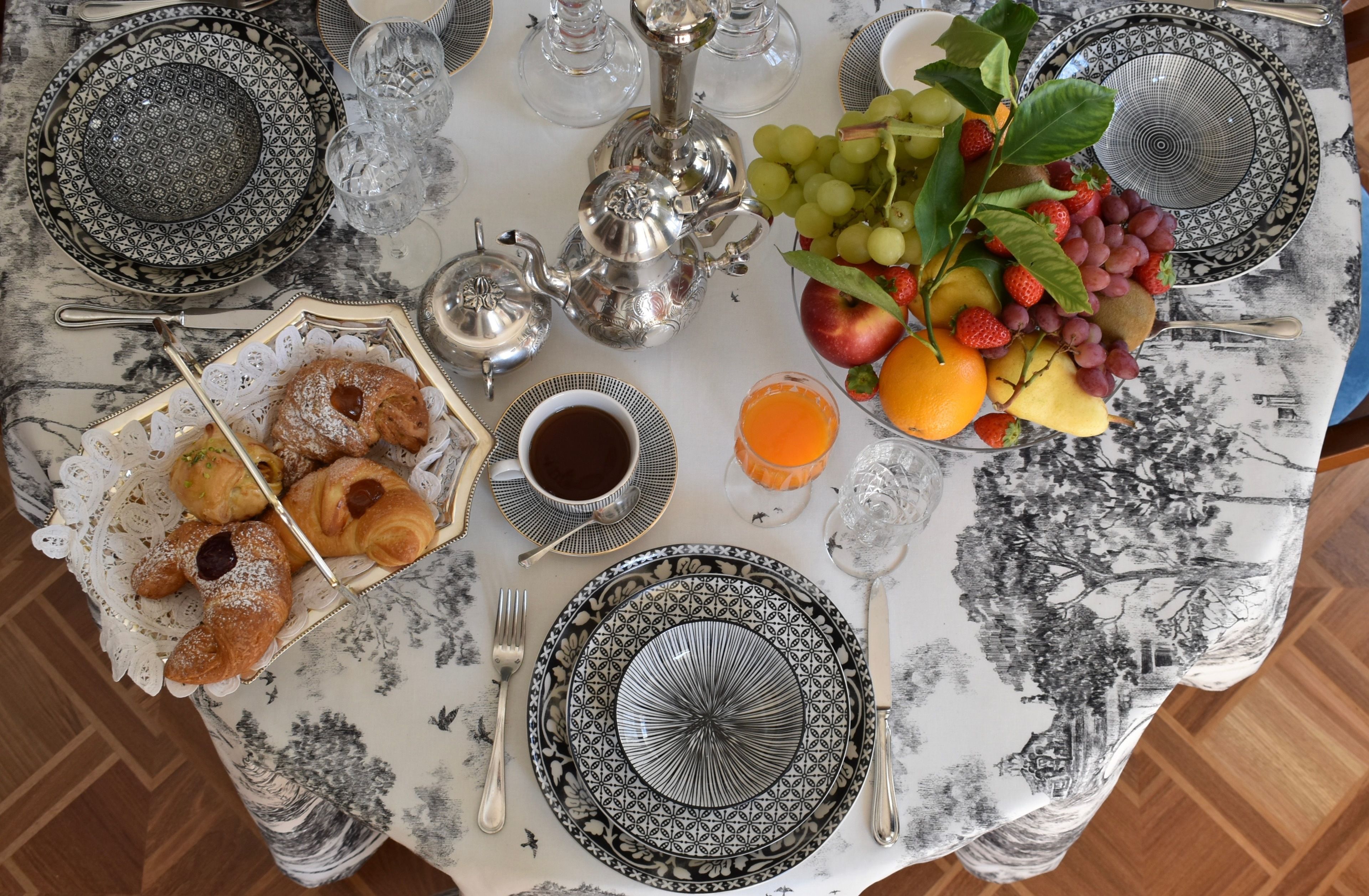Una mesa dispuesta con un mantel estampado, cubiertos, platos, una tetera, un frutero, pasteles y una taza de café.