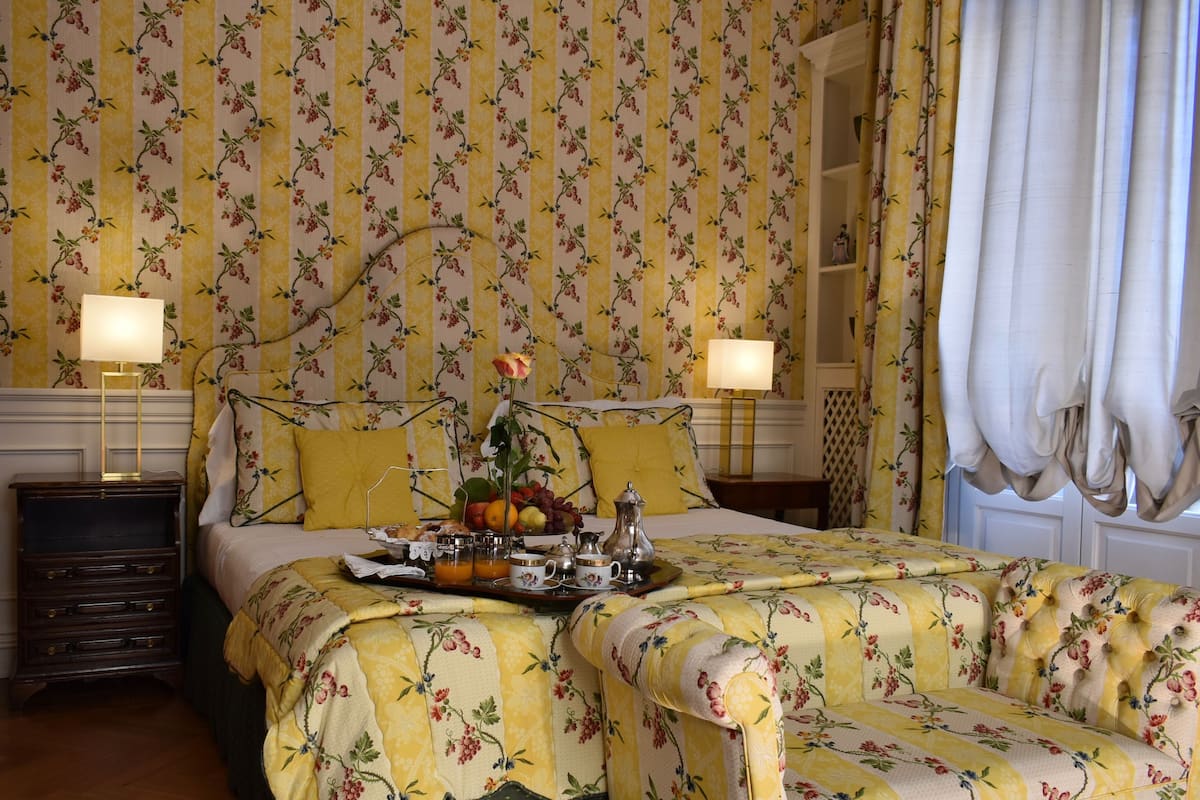 A hotel room with a floral wallpaper, a bed with a floral bedspread, two bedside lamps, a nightstand, and a window with curtains.