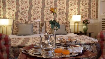 Ein ordentlich arrangiertes Frühstücksbrettchen auf einem Bett mit floralem Bettbezug, eine Blumenvase und eine Rosen-Zentralanordnung.