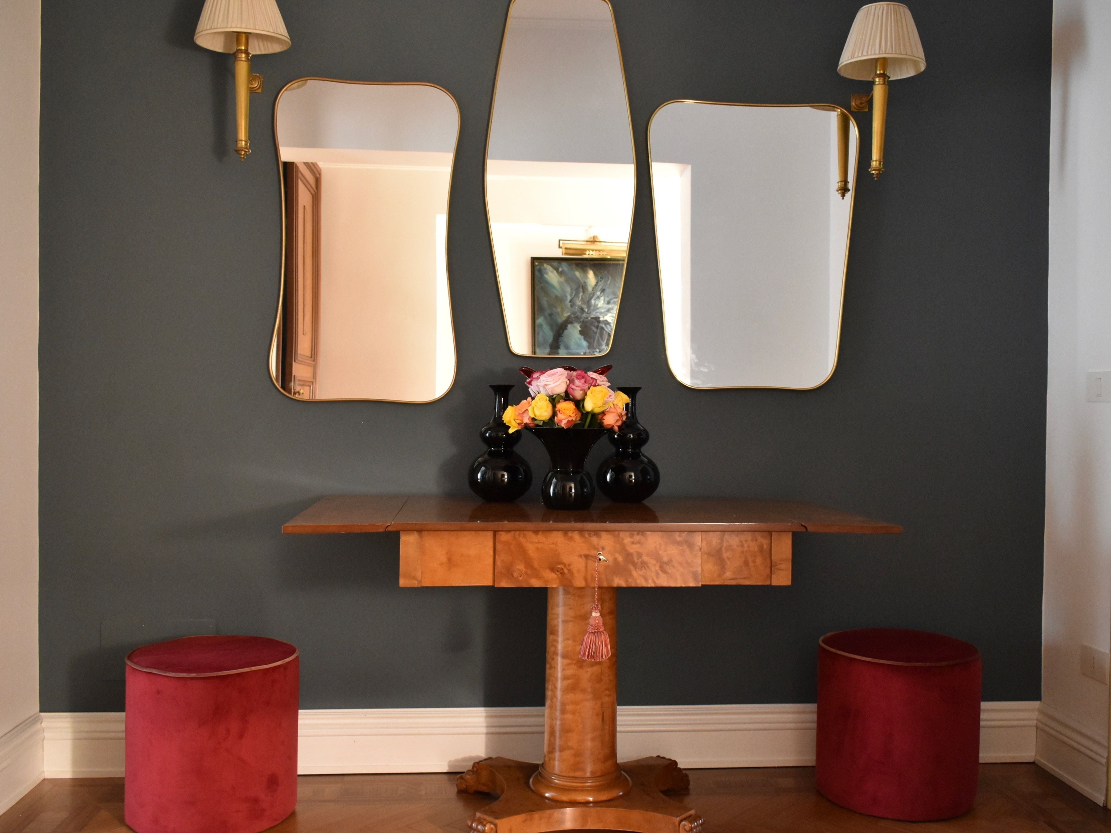 A wooden console table with a mirror and two black vases with flowers, flanked by red cylindrical stools, against a dark wall with gold-framed mirrors and wall sconces.
