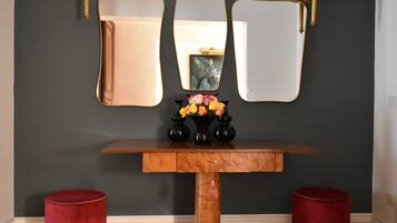A wooden console table with a mirror and two black vases with flowers, flanked by red cylindrical stools, against a dark wall with gold-framed mirrors and wall sconces.