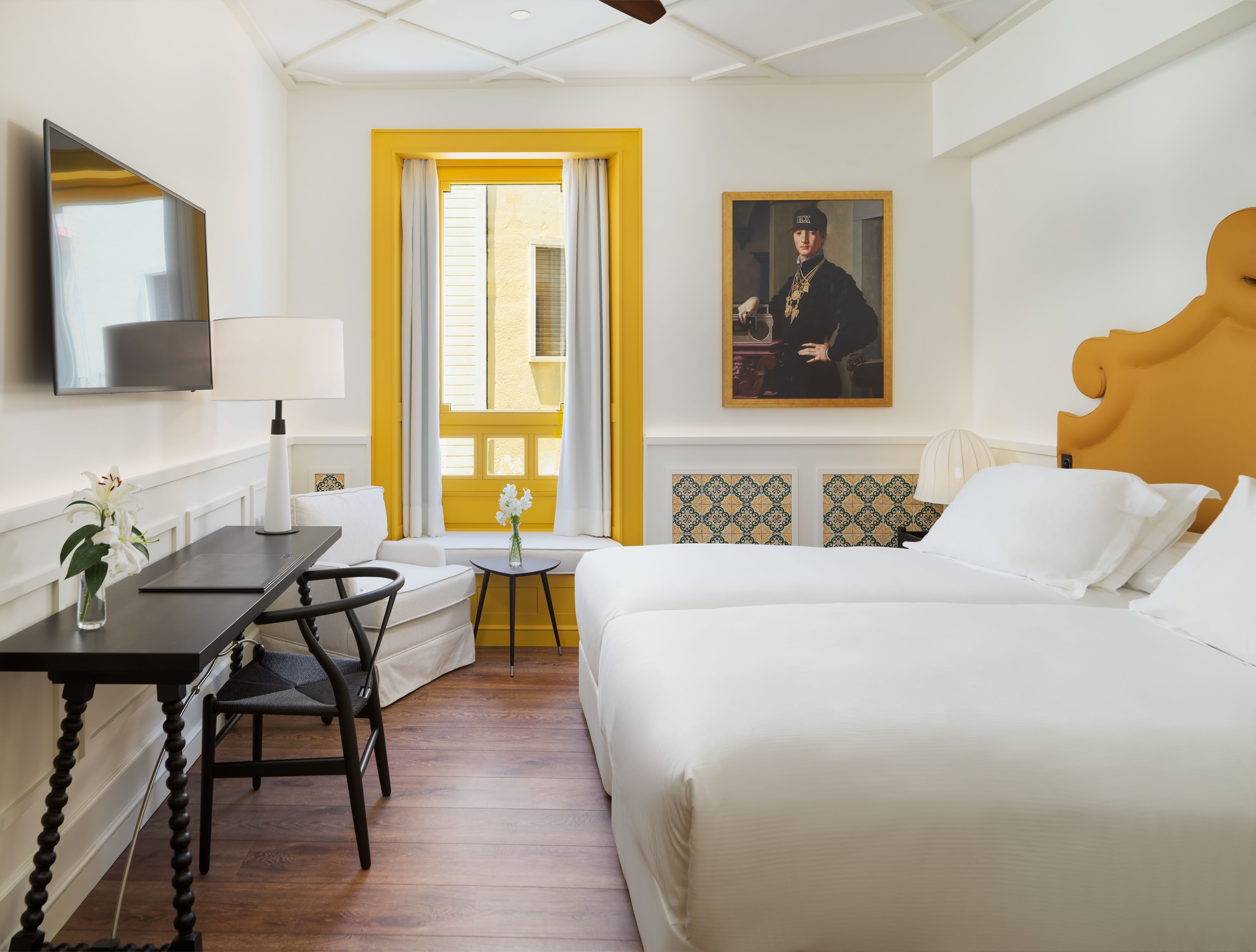A hotel room with a bed, a desk with a chair, a TV, a window with curtains, and a painting on the wall.
