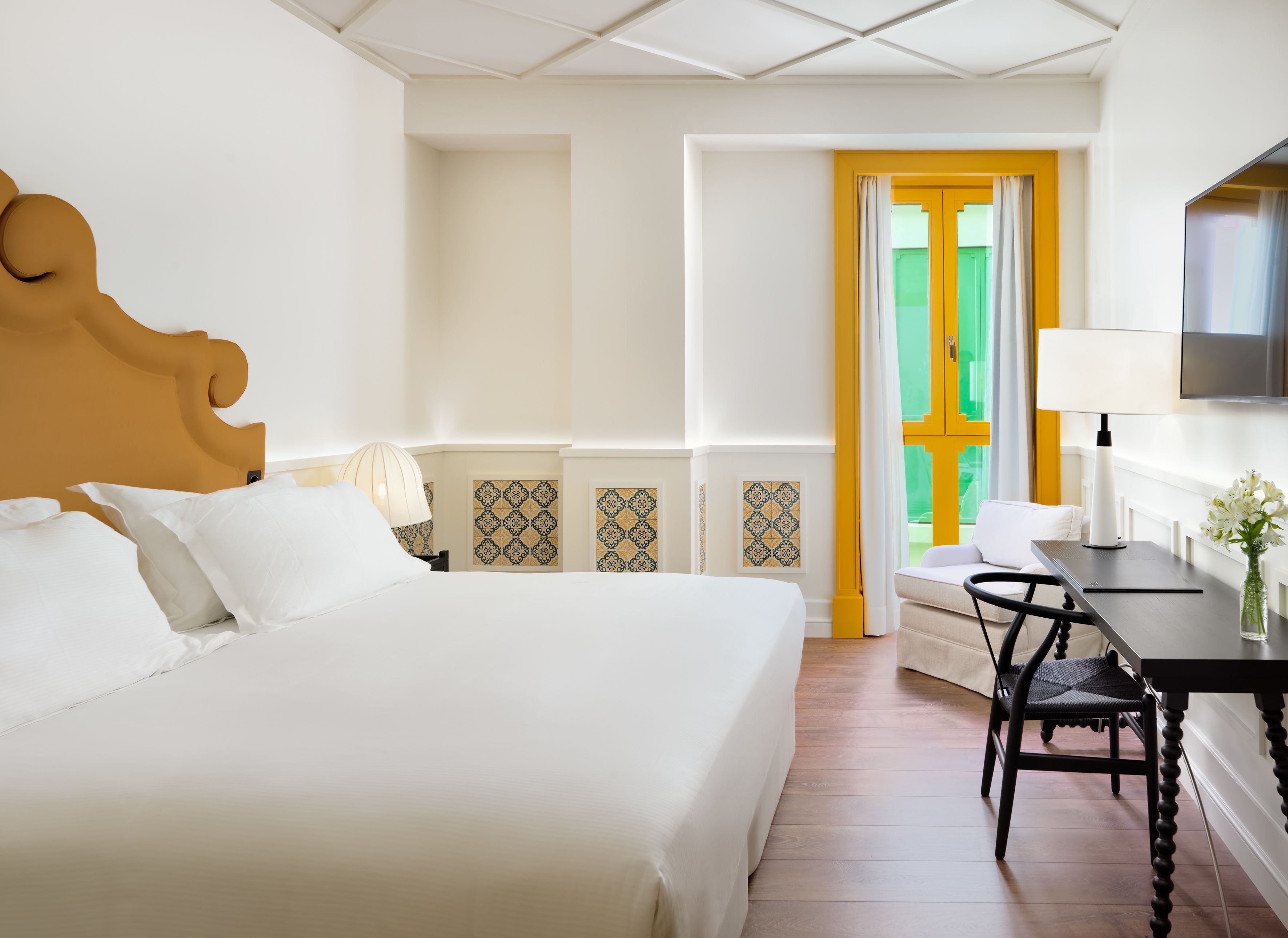 A hotel room with a large bed, a desk with a chair, a TV, and a window with a yellow door.
