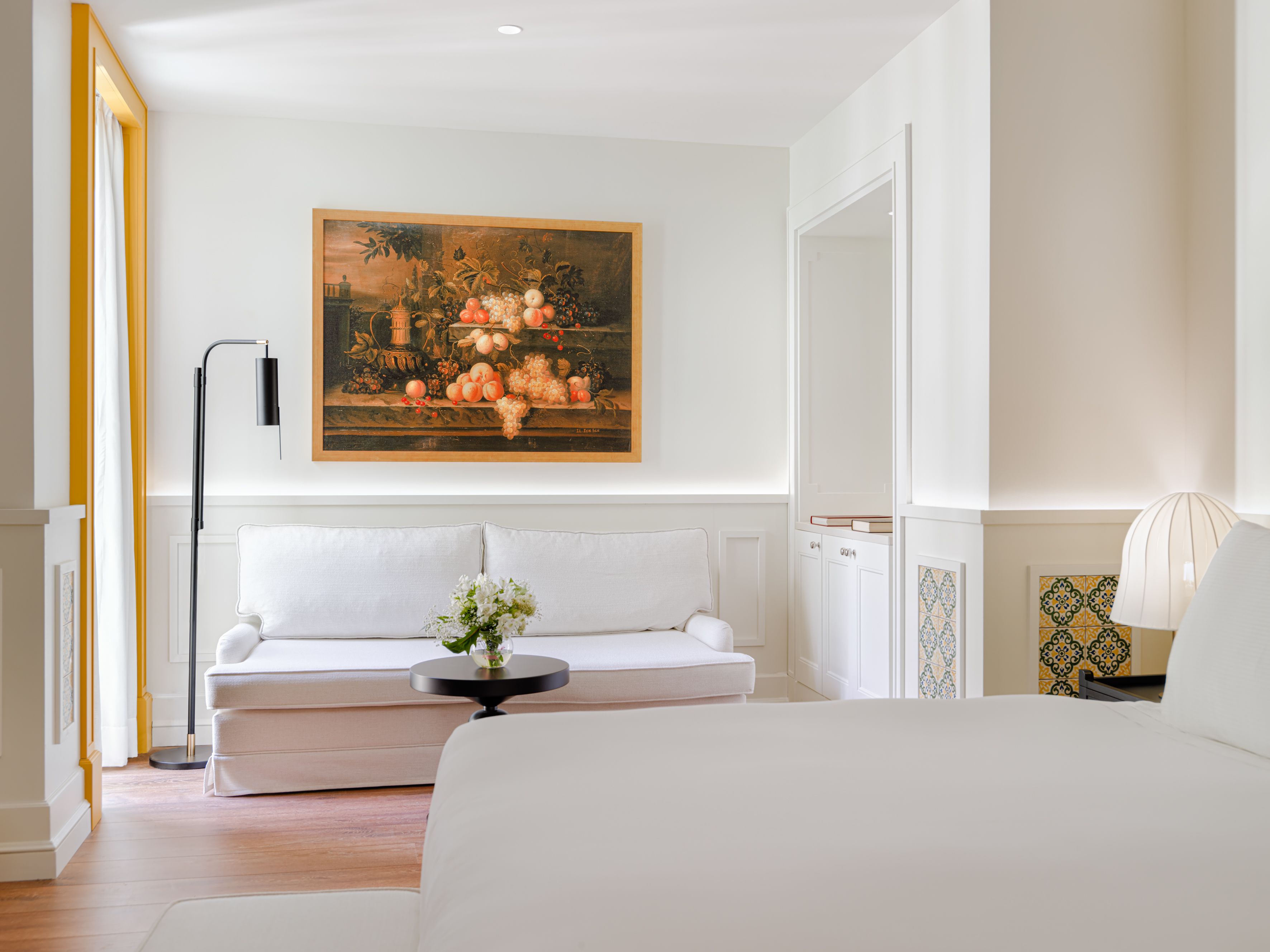 Un dormitorio moderno con un sofá blanco, una cama, un cuadro de frutas y una lámpara de pie.