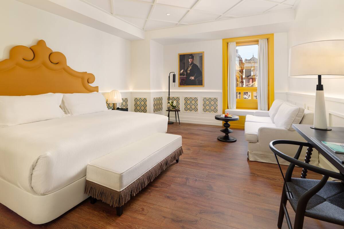 Une chambre d’hôtel avec un grand lit, un bureau, une chaise, un ventilateur de plafond et une fenêtre avec des rideaux.