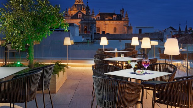 An outdoor dining area with wicker chairs and tables, a tree, and a view of a historic building at dusk. - H10 Casa de la Plata (Seville)