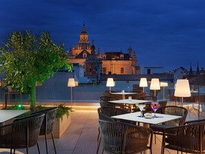 Un espacio al aire libre para comer con sillas y mesas de mimbre, un árbol y vistas a un edificio histórico al anochecer.