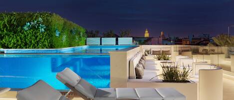 Un área de piscina en la azotea con sillones y vistas a un paisaje urbano al atardecer.