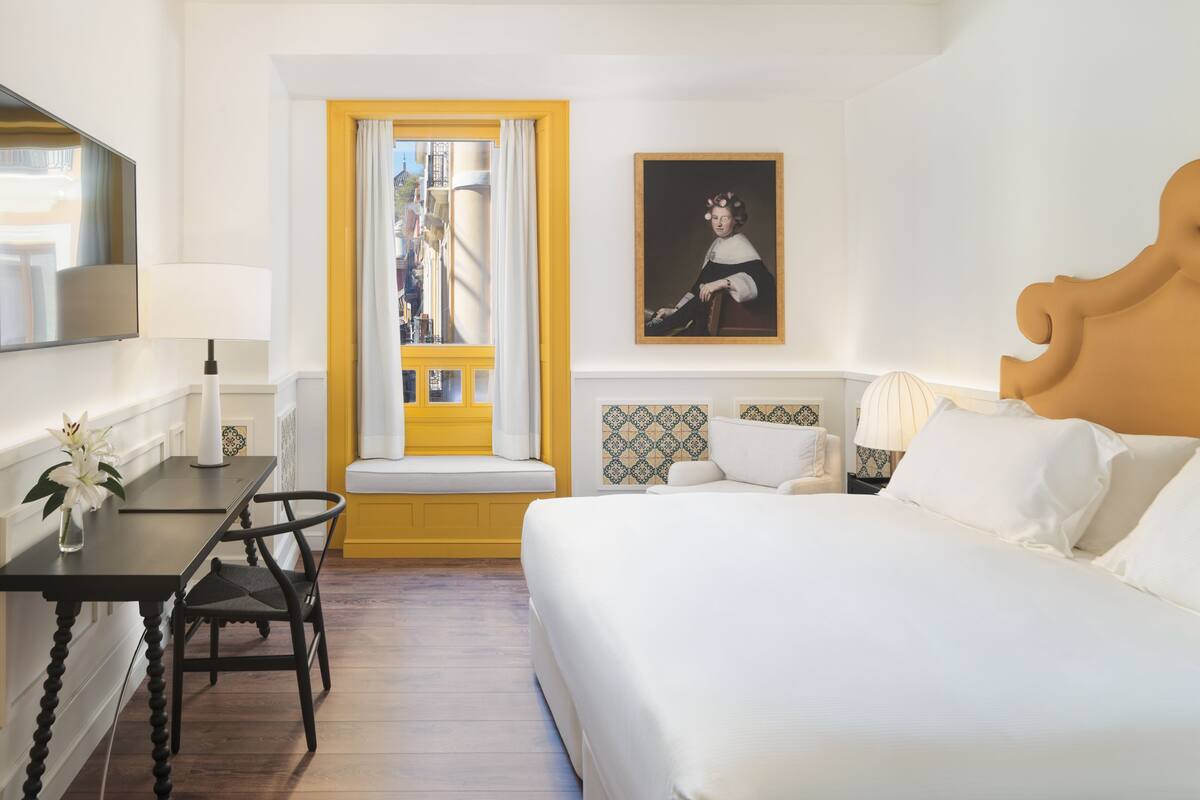 Une chambre d’hôtel avec un lit, un bureau, une chaise, une fenêtre avec des bordures jaunes et un tableau accroché au mur.