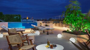 A rooftop pool area with lounge chairs, a table with drinks, and a view of the city skyline at dusk. - H10 Casa de la Plata (Seville)