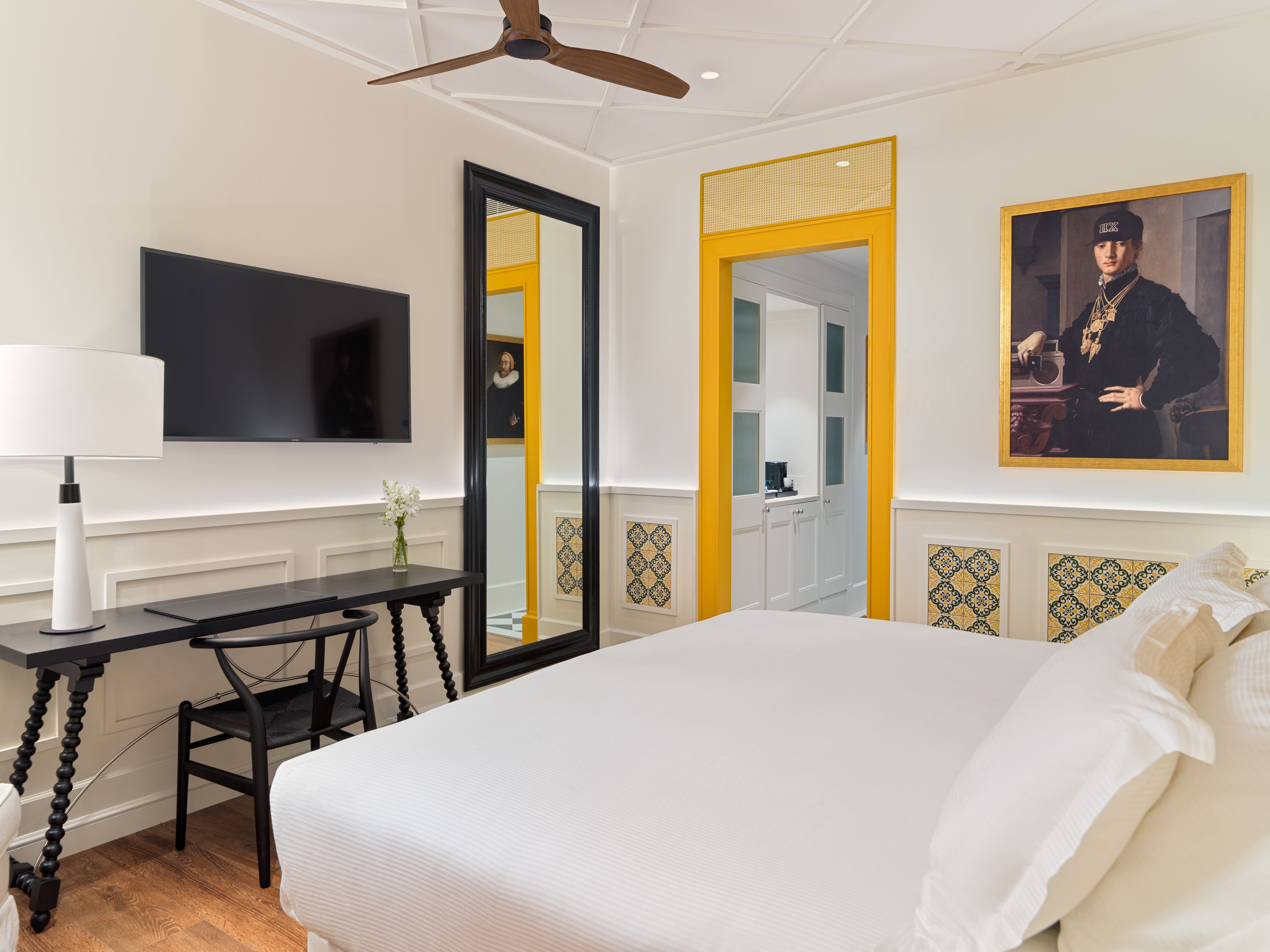 A hotel room with a large bed, a desk with a lamp, a flat-screen TV, and a ceiling fan.