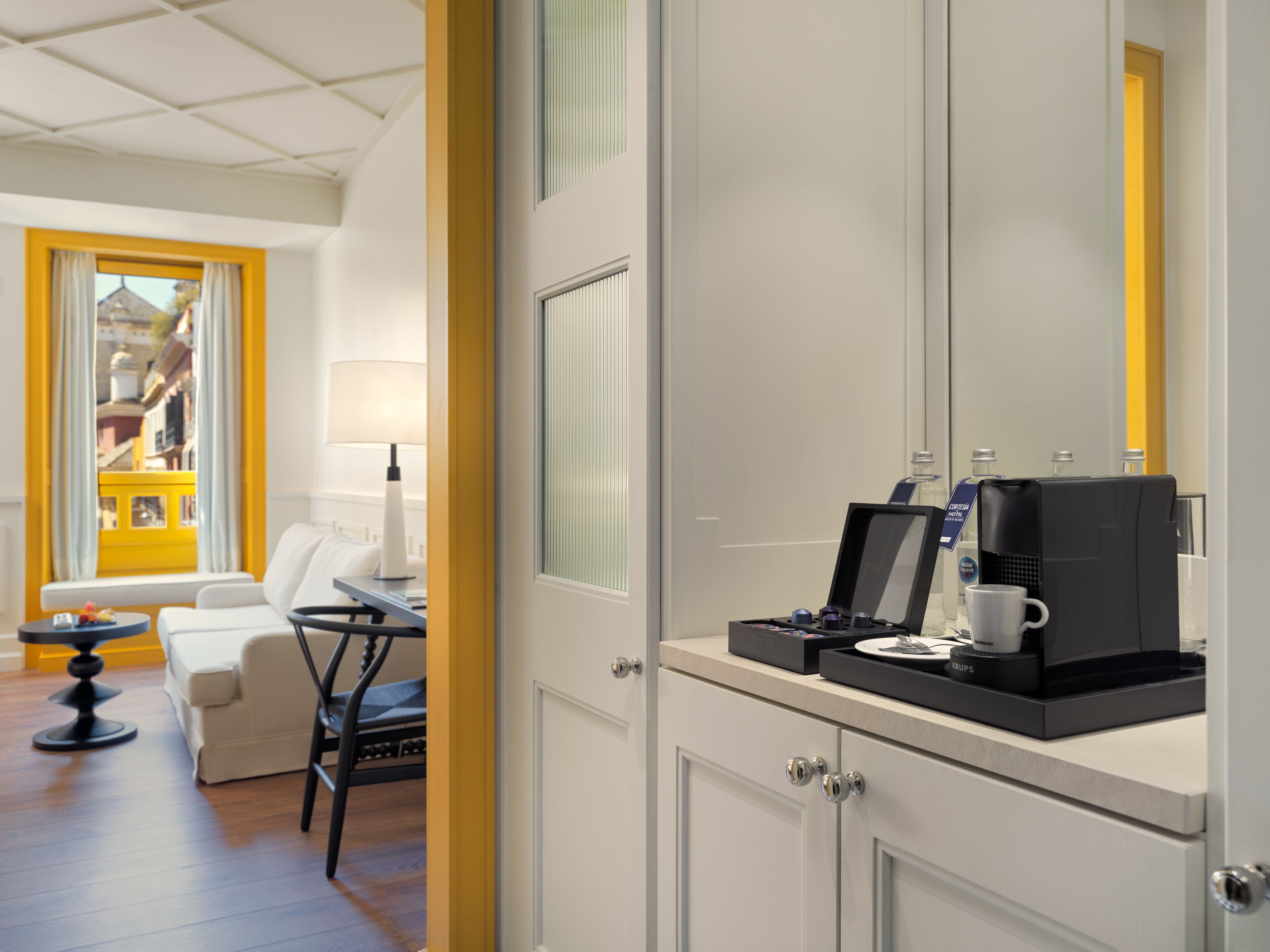 Ein Hotelzimmer mit Bett, Schreibtisch mit Kaffeemaschine, Stuhl, Lampe und Blick auf ein gelbes Gebäude durch ein Fenster.