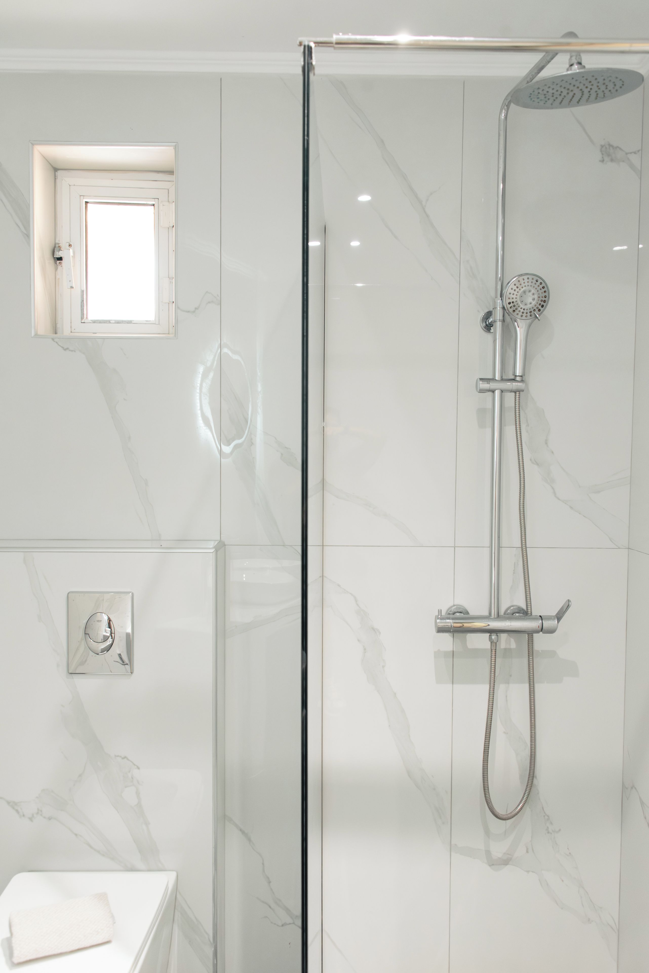 Un baño moderno con ducha acristalada, un ventanal pequeño y una toalla sobre una encimera blanca.