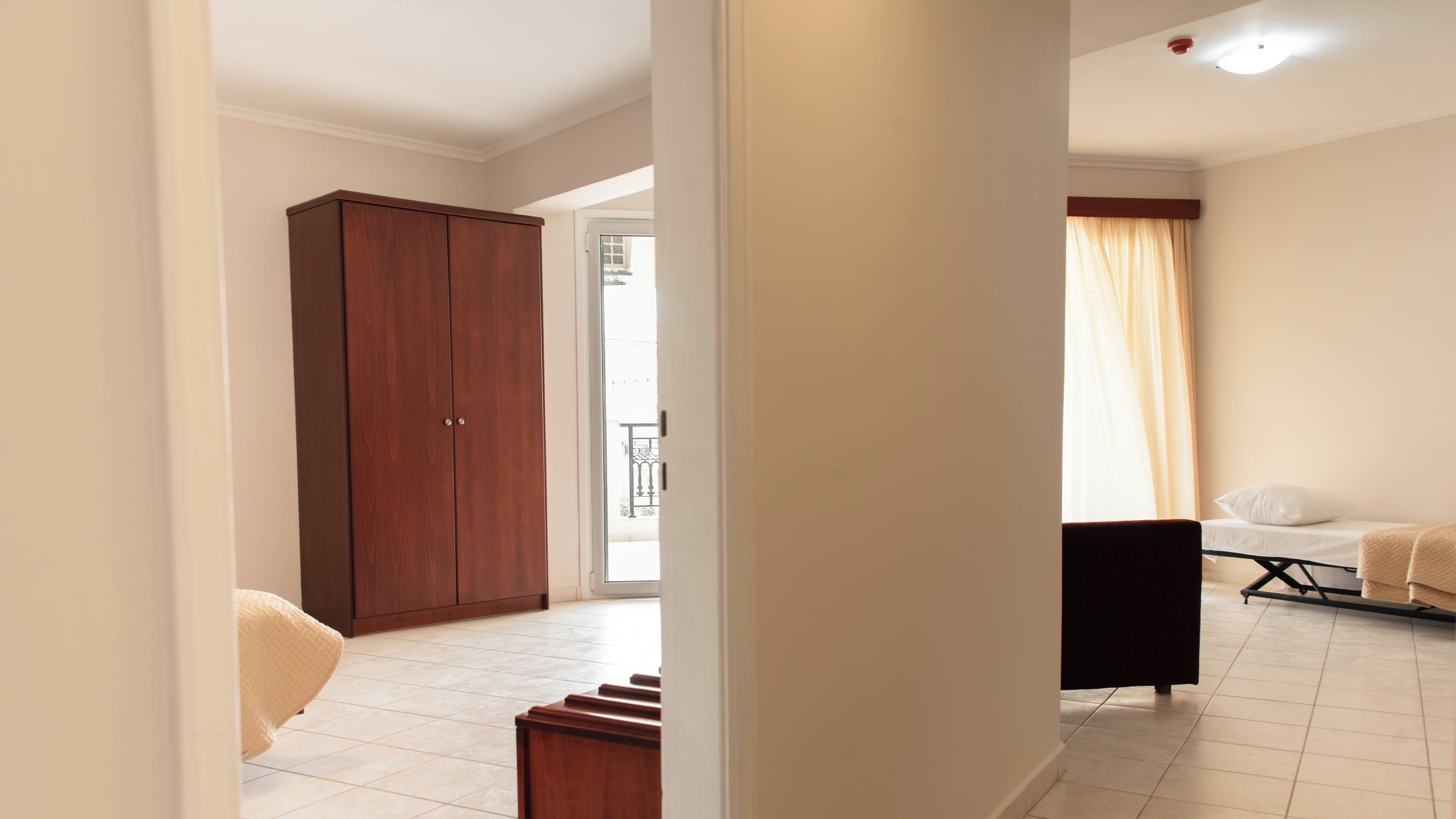 A hotel room with a large wardrobe, a bed with a white pillow, a small table, and a window with curtains.