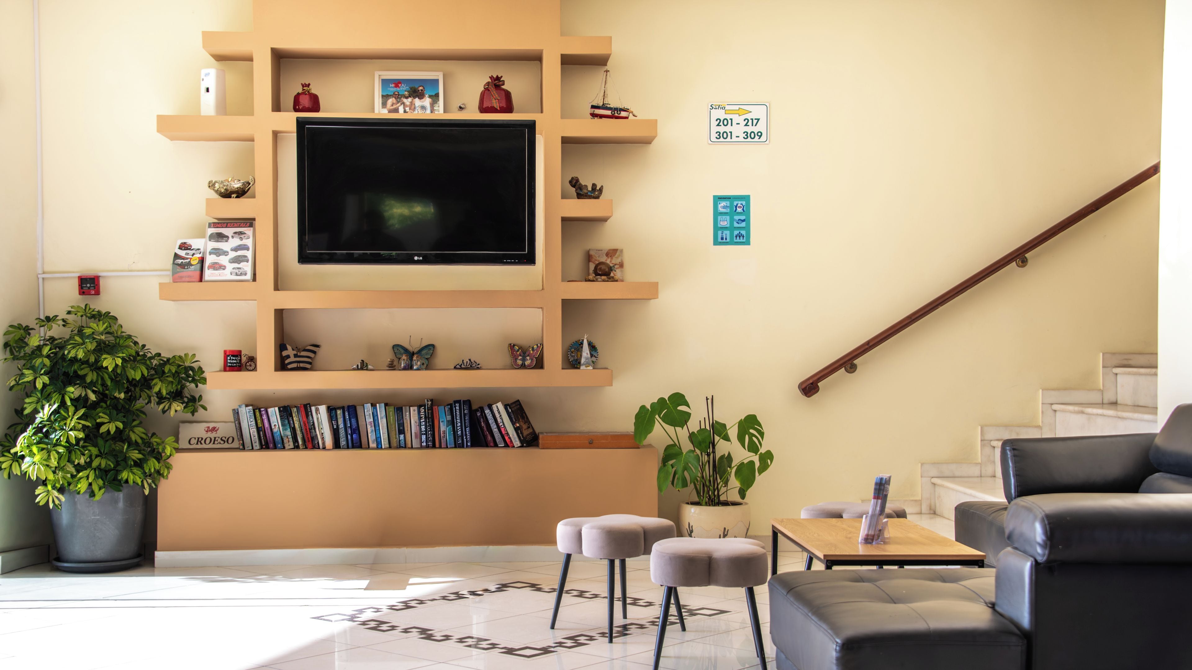 A hotel lobby with a flat-screen TV, bookshelves, a potted plant, a staircase, and a leather sofa.