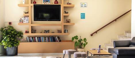 A hotel lobby with a flat-screen TV, bookshelves, a potted plant, a staircase, and a leather sofa.