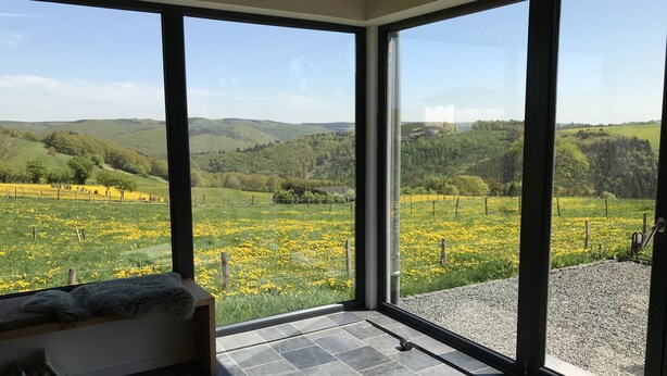 Una habitación con grandes ventanales que ofrecen vista a un campo verde con flores amarillas y colinas onduladas.