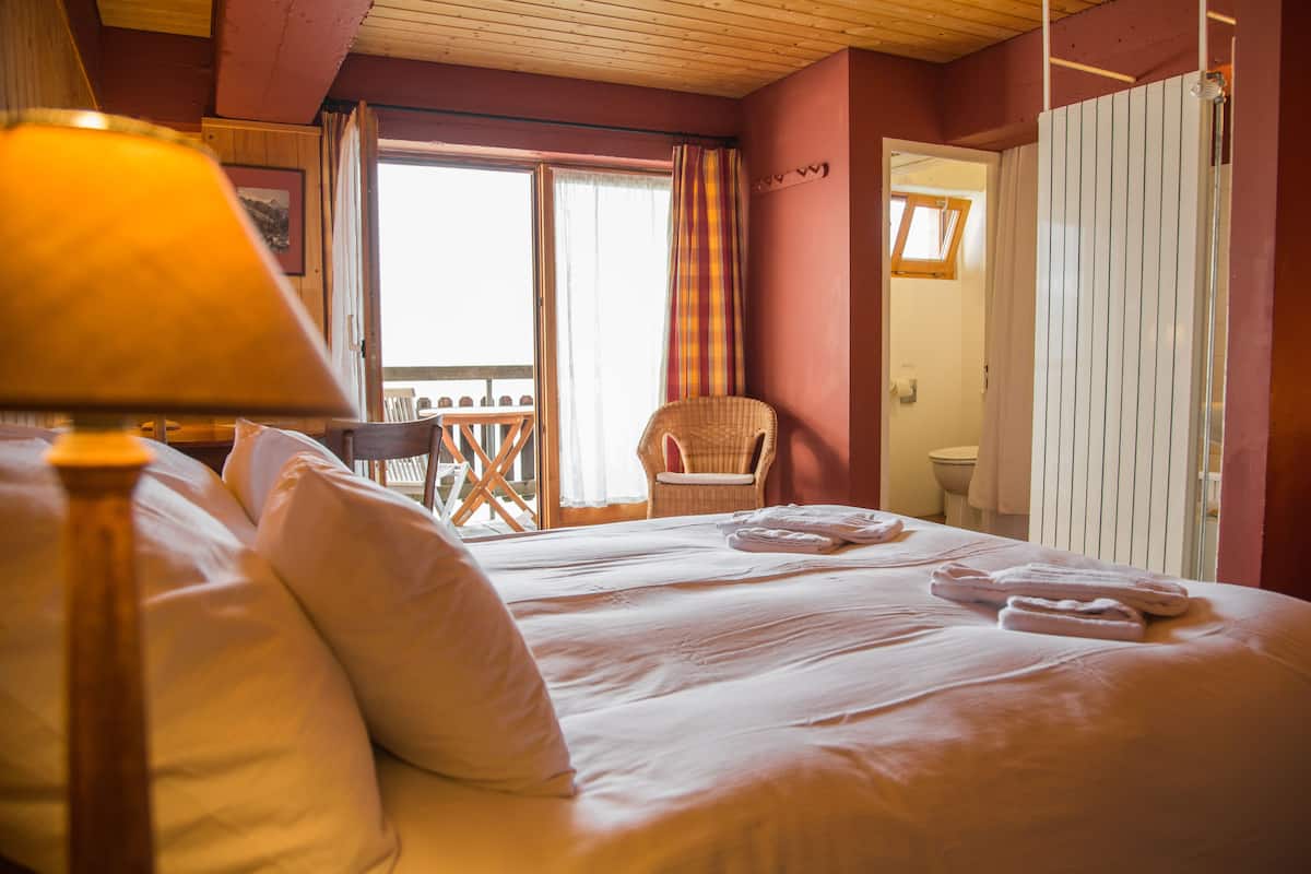 A hotel room with a bed, pillows, a chair, a table, and a window with curtains.
