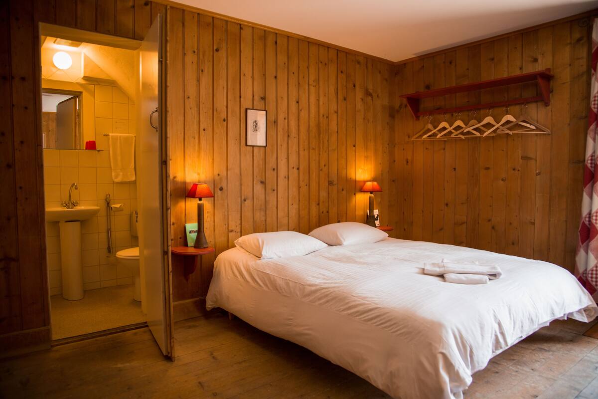 A wooden-paneled room with a bed, a nightstand, a mirror, and a hanging clothes rack.