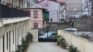 Um beco estreito com vasos de plantas ao longo das laterais, um portão de entrada e edifícios ao fundo.