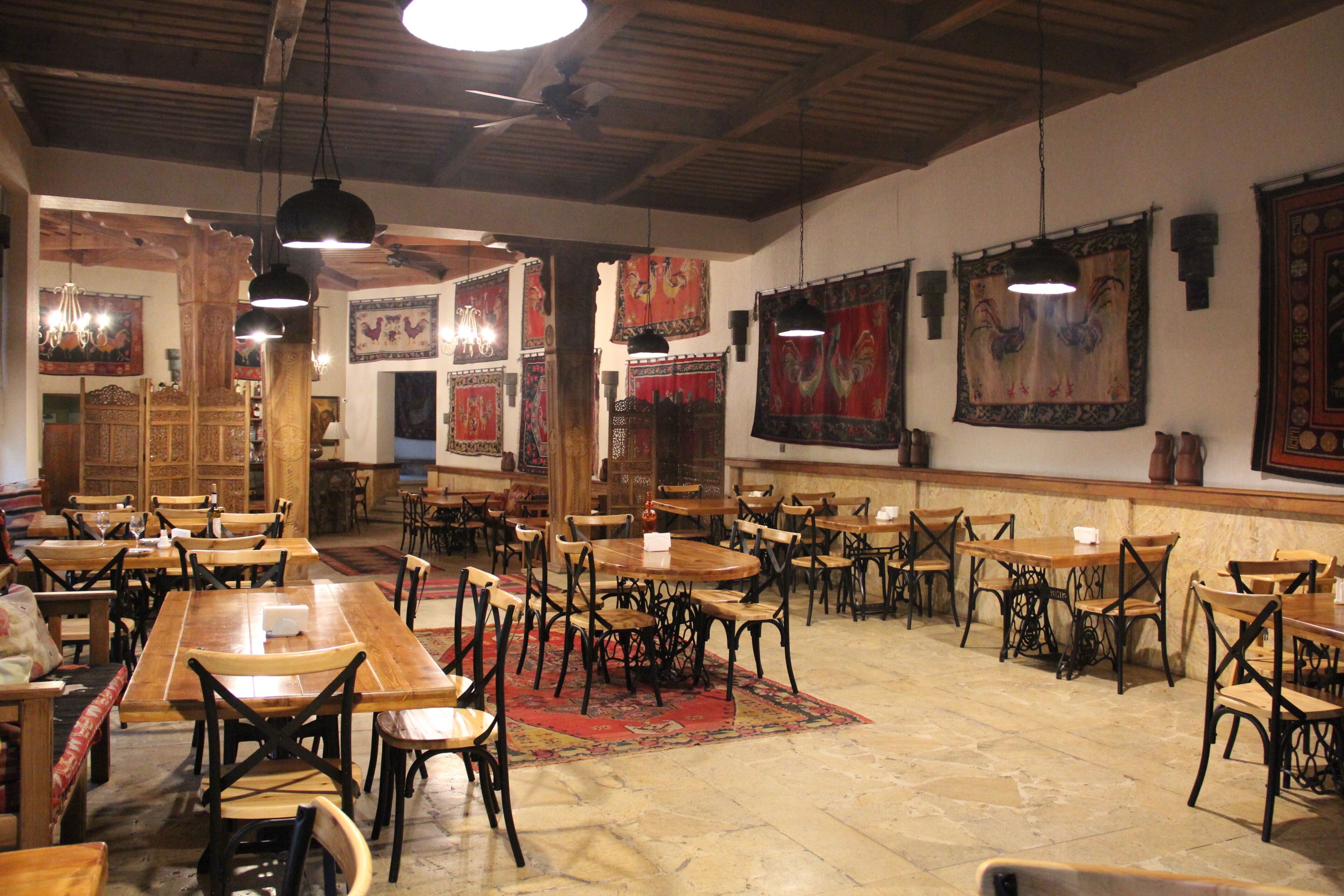 A restaurant interior with wooden tables and chairs, hanging lights, and decorative tapestries on the walls.