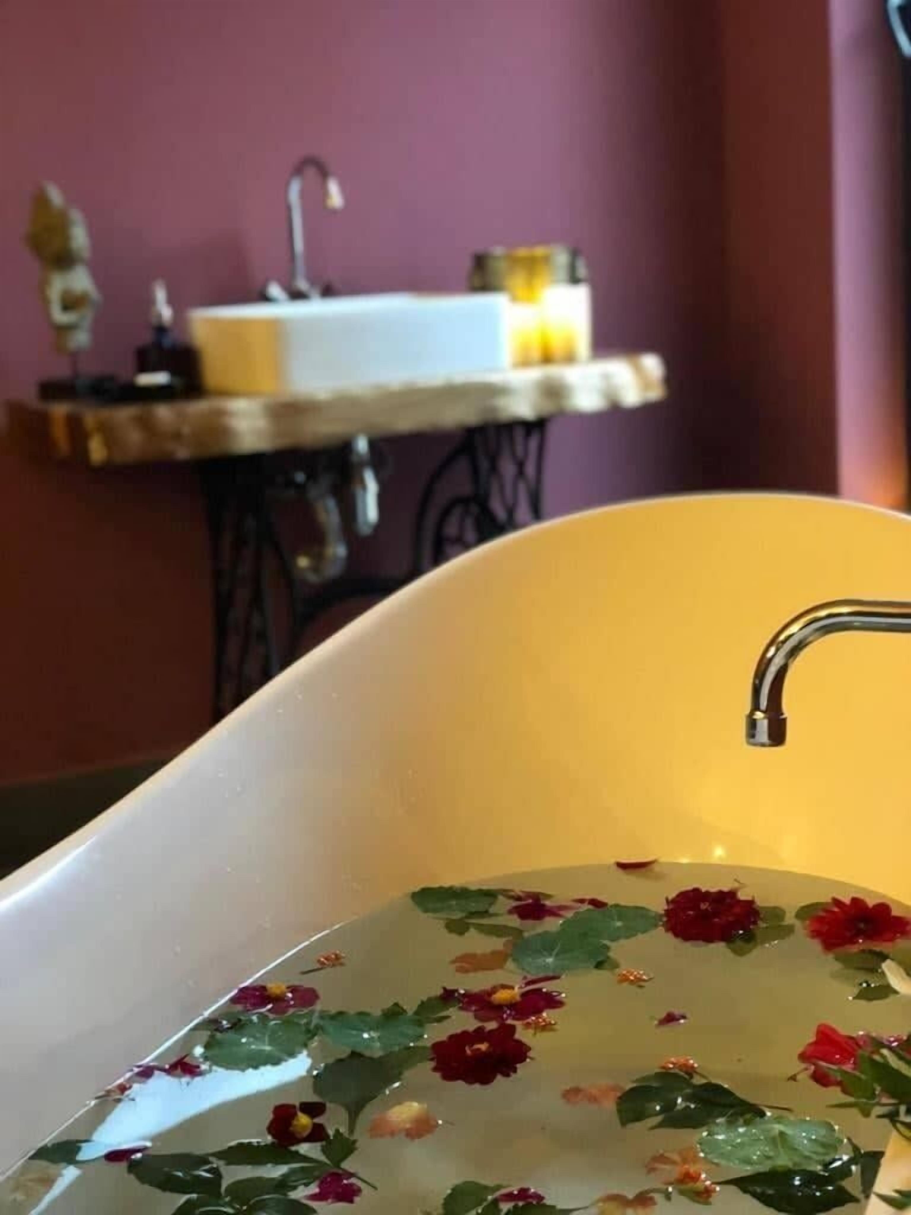 A bathtub with floral decorations in water, a sink, and a small table with a lamp in a room with purple walls.