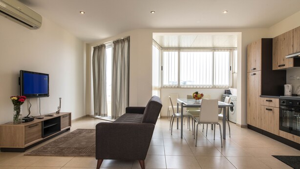 A modern living room with a sofa, a TV, a dining table with chairs, and a kitchen area with cabinets and appliances. - Vacation Rentals (St. Paul's Bay)