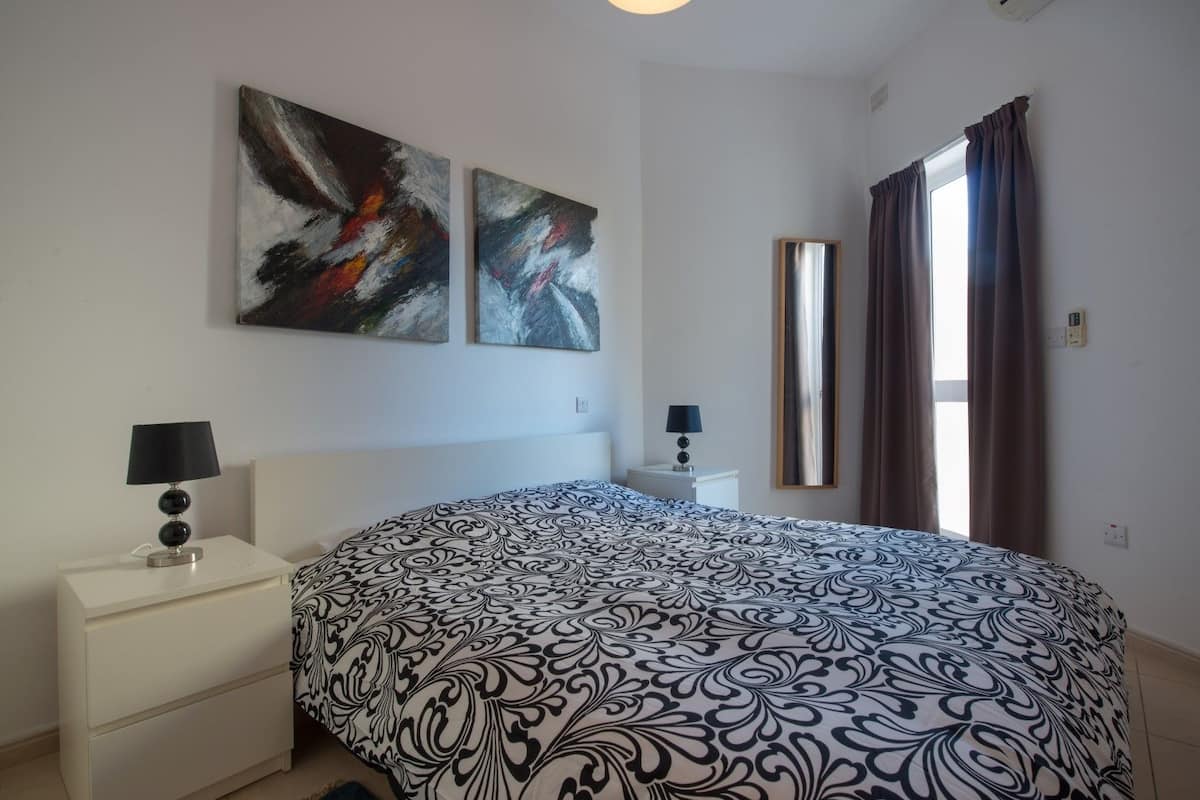 A bedroom with a large bed, two bedside tables with lamps, a mirror, and abstract paintings on the wall.
