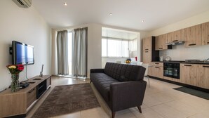 A modern living room with a sofa, TV, and entertainment unit. A kitchen with wooden cabinets and appliances is visible in the background. - Vacation Rentals (St. Paul's Bay)