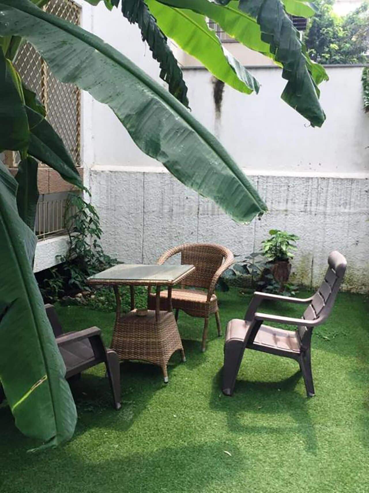 Une petite terrasse extérieure avec une table et deux chaises en osier, entourée d’une végétation luxuriante et d’un mur blanc.