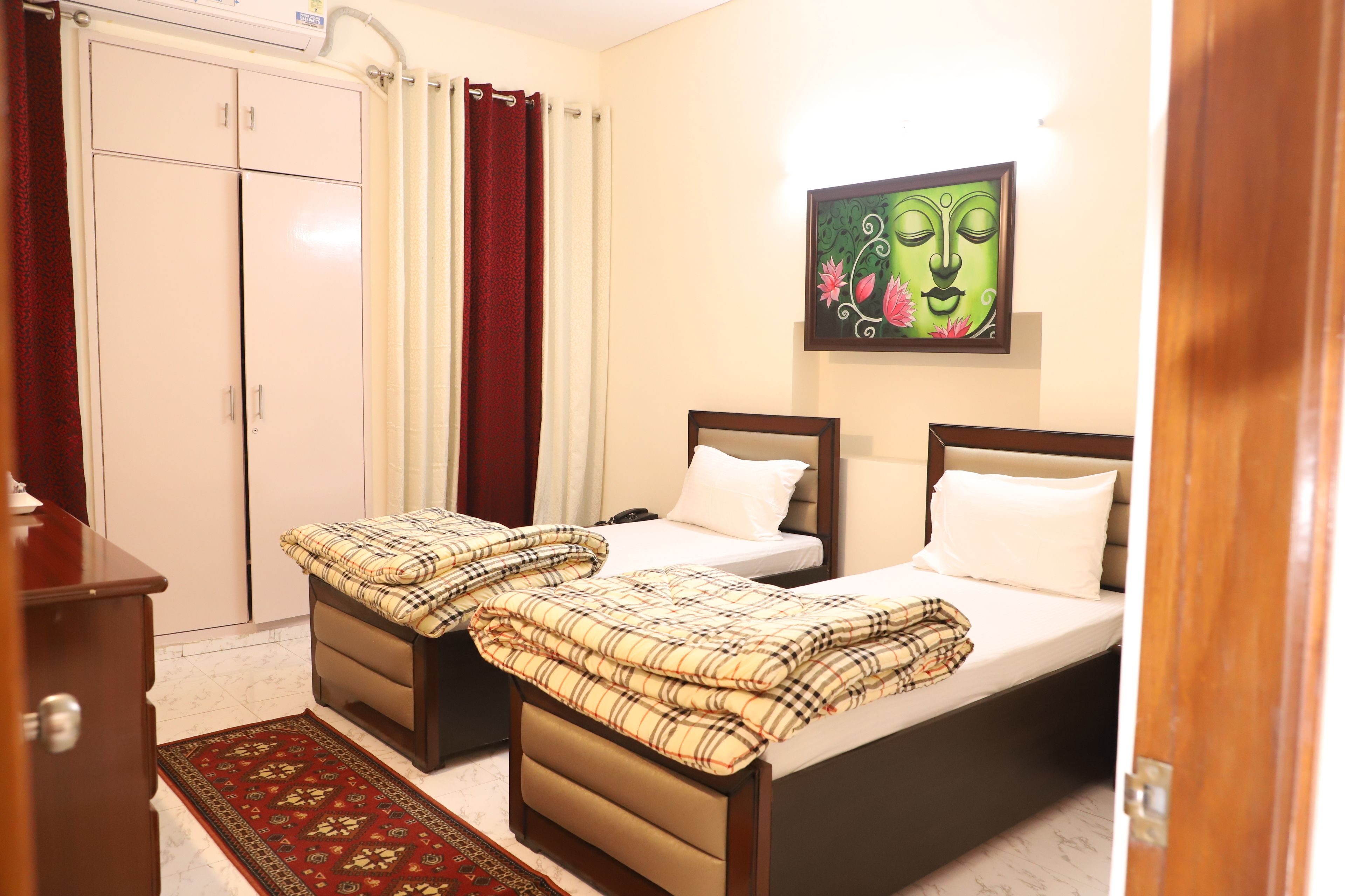 A hotel room with two beds, a wardrobe, a red patterned rug, and a painting on the wall.