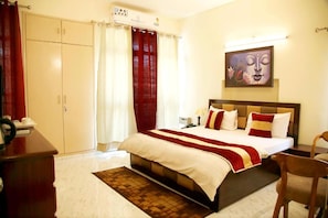 A hotel room with a bed, a desk, a chair, a wardrobe, and a painting on the wall. - Maplewood Stay (New Delhi)