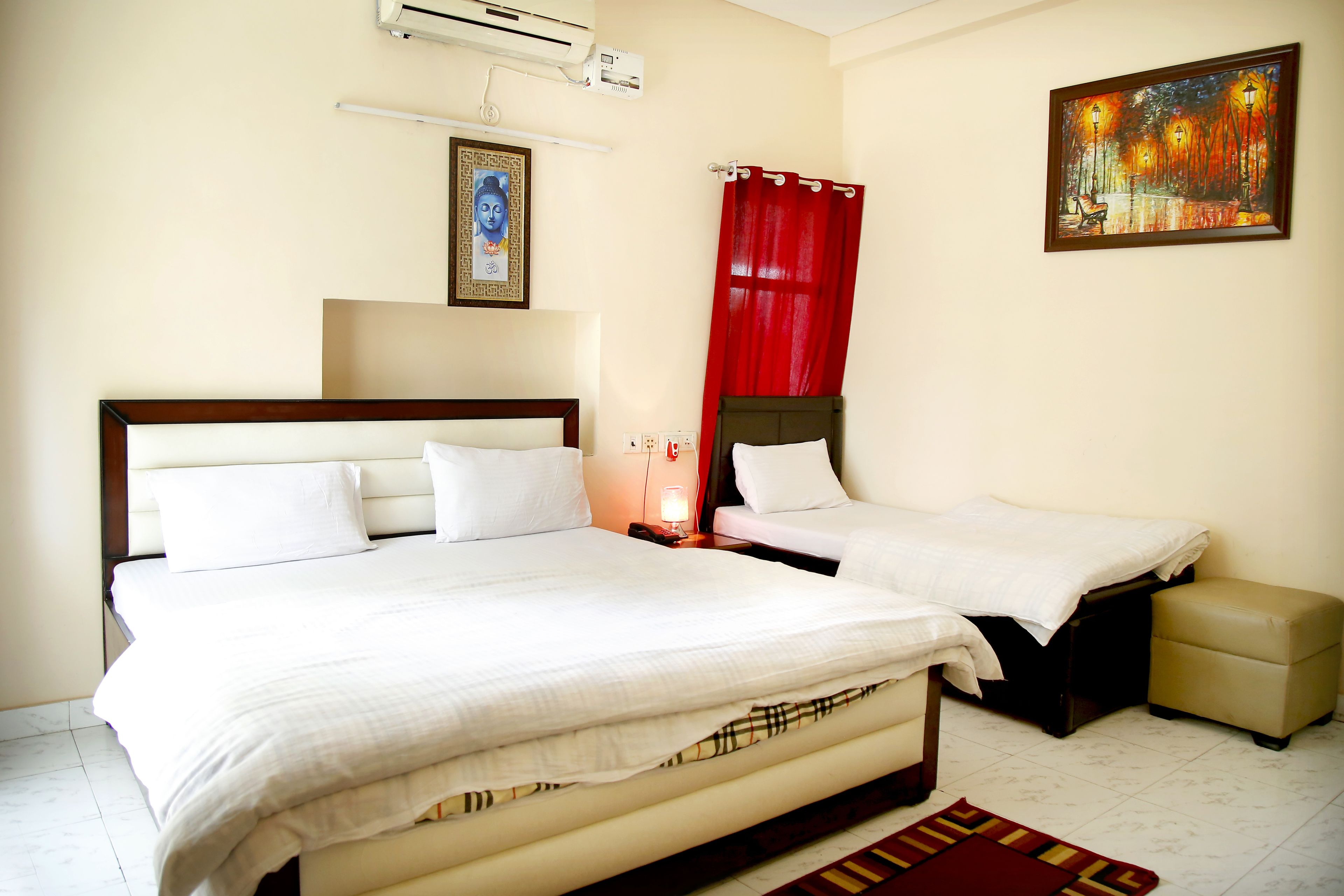 A hotel room with a large bed, a smaller bed, a red curtain, a painting, and a small table with a lamp.