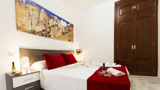 A hotel room with a bed, a nightstand, a red throw blanket, a bottle of wine, and a painting of a mountain landscape. - Apartamentos El Tajo (Ronda)