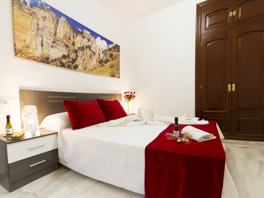 Habitación de hotel con cama, mesita de noche, una manta roja, una botella de vino y un cuadro de un paisaje montañoso.