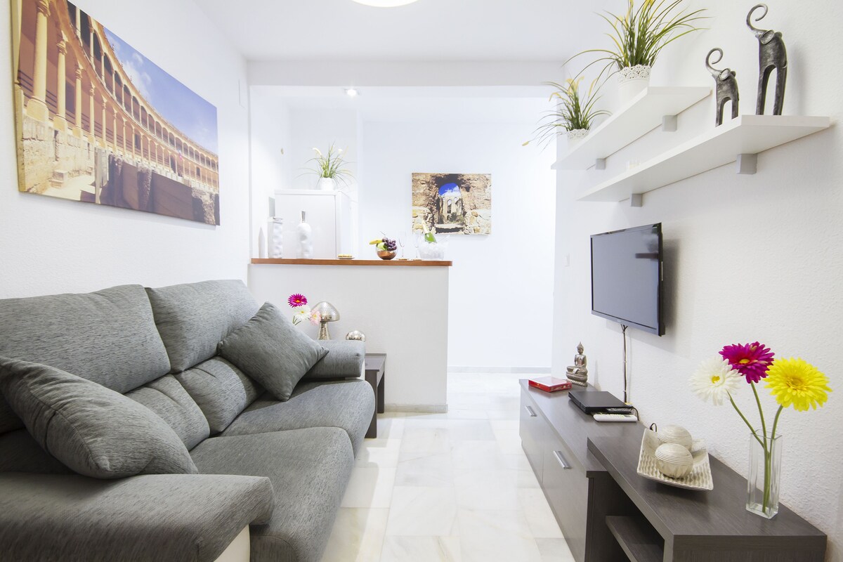 En moderne stue med en grå sofa, et fladskærms-tv og dekorative elementer, herunder blomster og figurer.