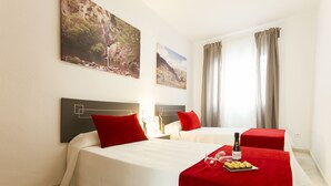 A hotel room with two beds, red pillows, a table with a bottle and snacks, and two landscape paintings on the wall. - Apartamentos El Tajo (Ronda)