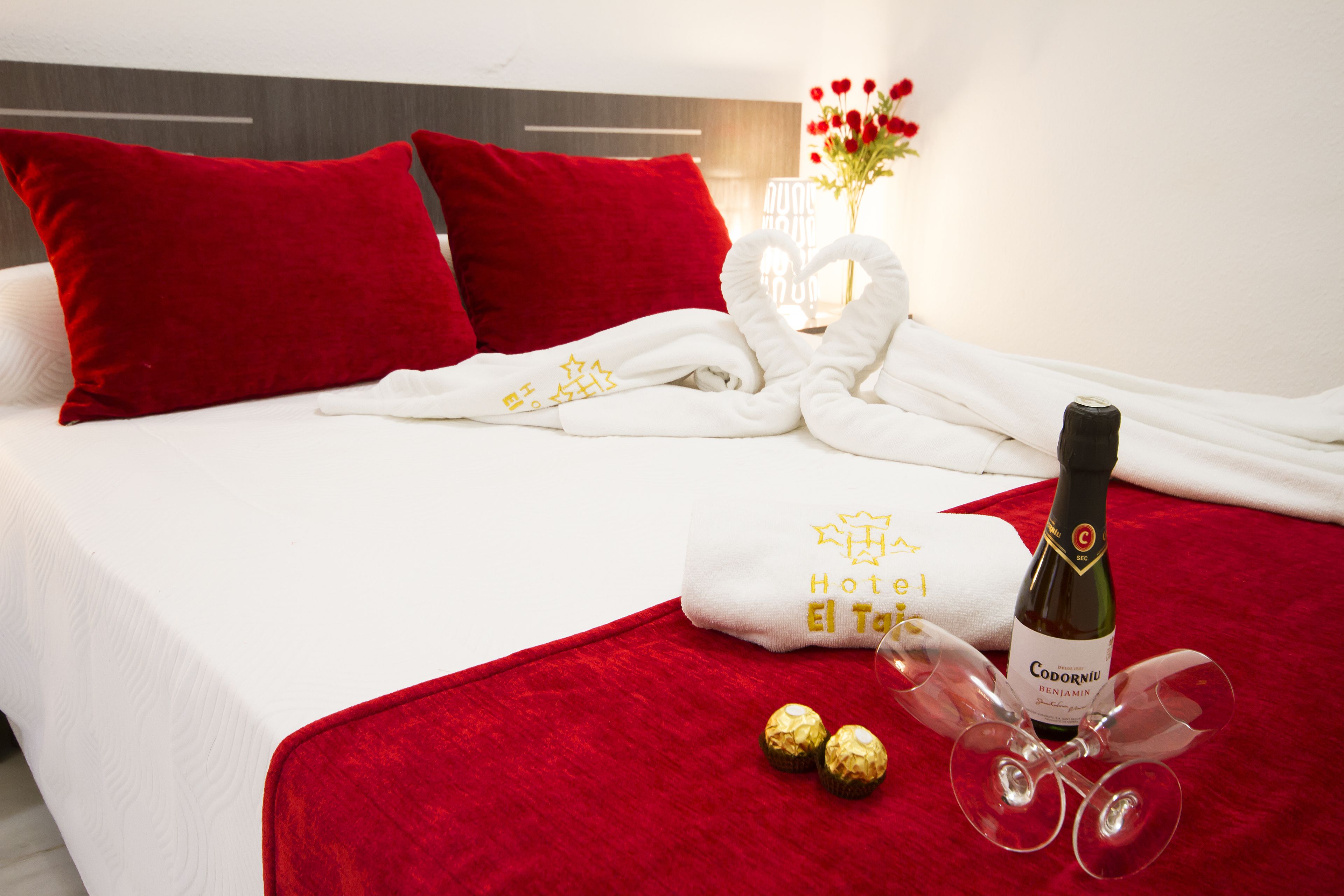 A neatly made bed with red pillows, a white towel, a bottle of wine, two glasses, and two chocolates.