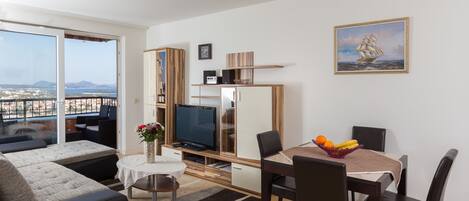A modern living room with a TV, dining table, and chairs. There is a balcony with a view of the city and mountains.