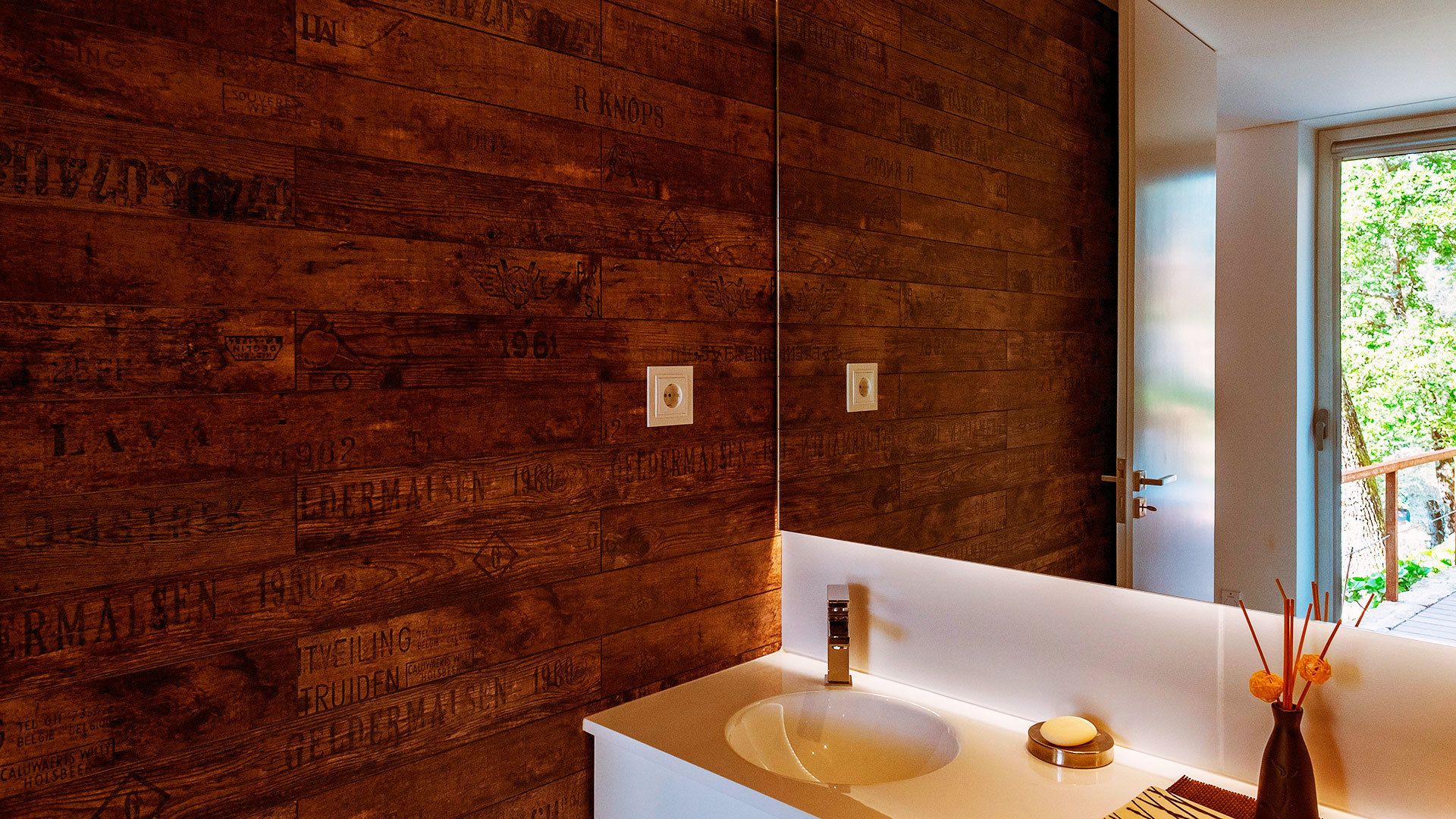 A bathroom with a wooden wall, a white sink, and a mirror reflecting a door and greenery outside.