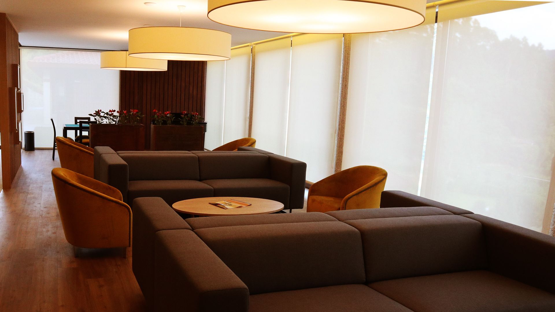 A modern living room with a sectional sofa, a round coffee table, and two armchairs. There are large windows with sheer curtains, a wooden floor, and a dining area in the background.