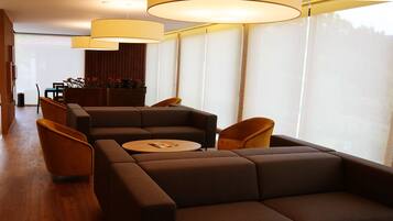 A modern living room with a sectional sofa, a round coffee table, and two armchairs. There are large windows with sheer curtains, a wooden floor, and a dining area in the background.