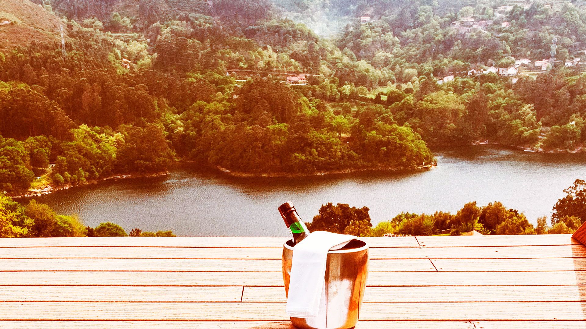 Una botella de champán en un balde de metal sobre una cubierta de madera con vista a un lago rodeado de árboles.