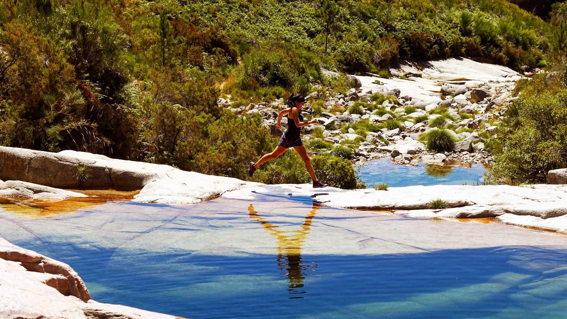 A person running on a rocky path with a clear pool of water in the foreground.