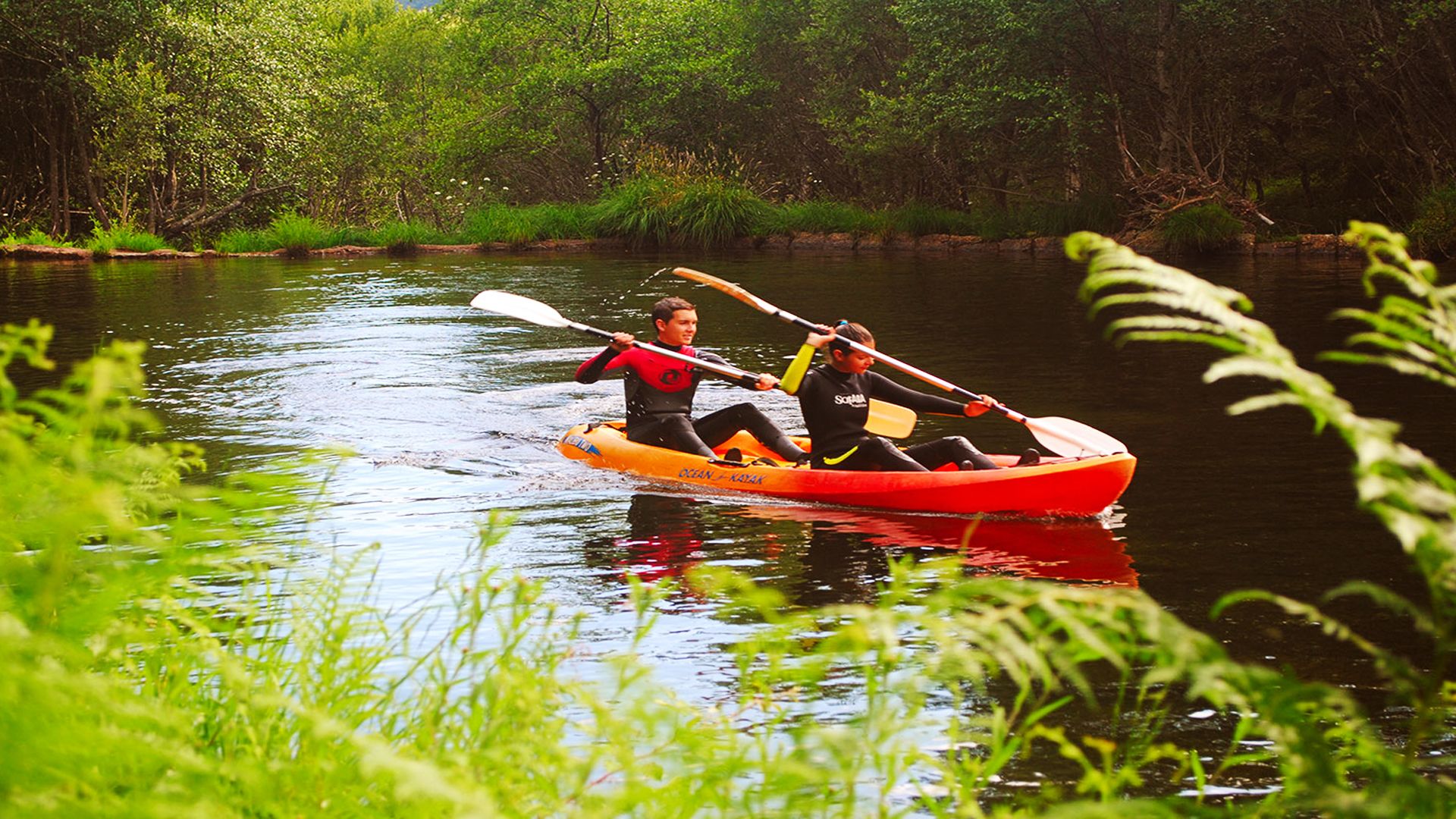 Two people kayaking in a narrow river surrounded by lush greenery.