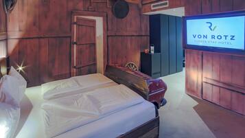 A wooden-themed hotel room with a bed, a vintage car, and a TV displaying the hotel name.