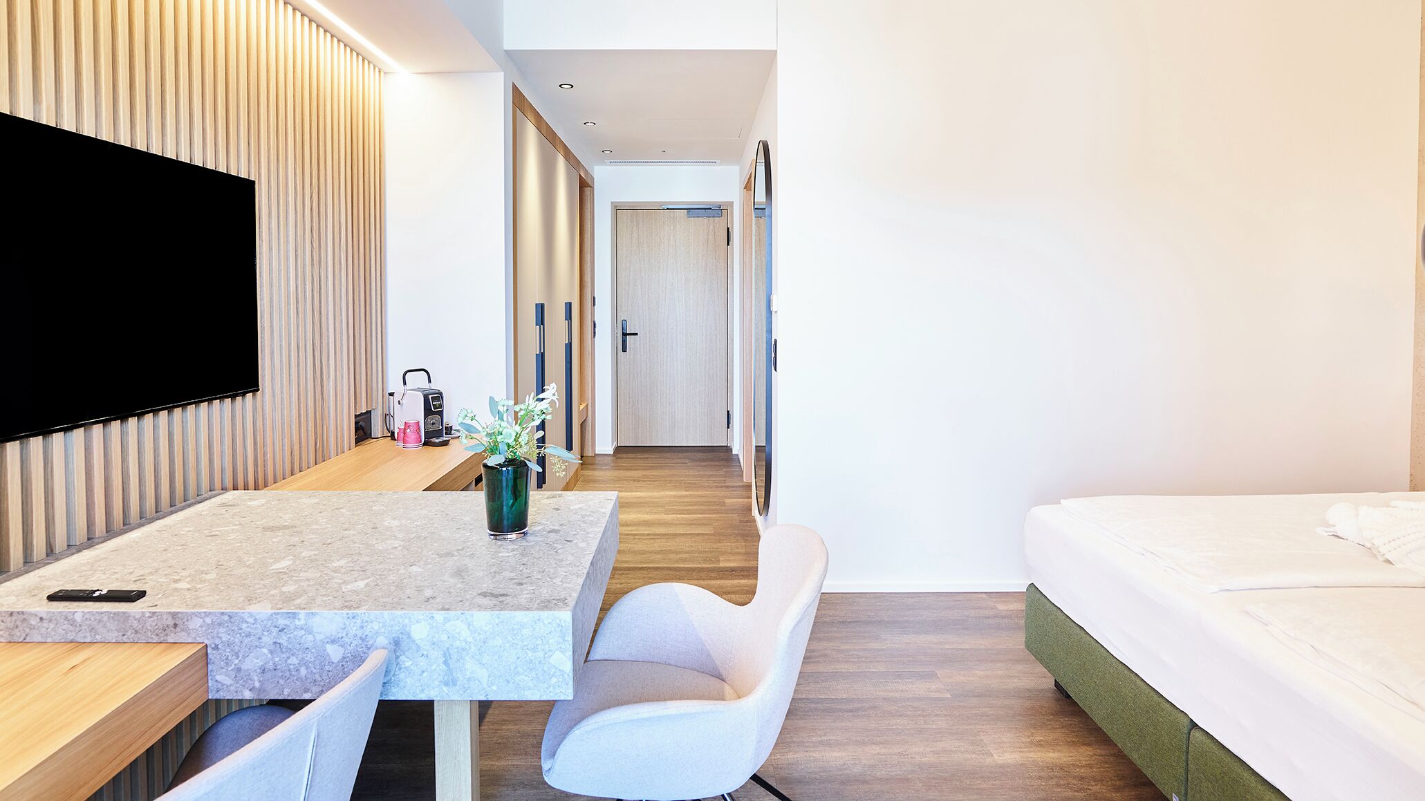 Una habitación de hotel moderna con una televisión de pantalla plana, un escritorio de madera, una silla blanca, una cama y un baño visible a través de una puerta.