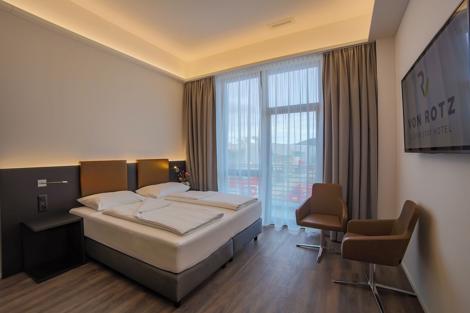 Habitació d'hotel amb un llit gran, dues taules de nit, un escriptori, una cadira i una finestra ampla amb cortines.