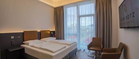 A hotel room with a large bed, two bedside tables, a desk, a chair, and a large window with curtains.