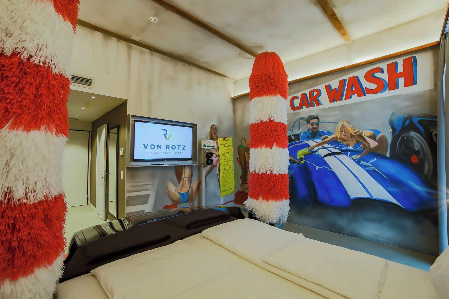 Ein Hotelzimmer mit Bett, einem Fernseher, der das Logo des Clever Stay Hotels zeigt, einer Wandmalerei mit einer Autowaschanlage und einer roten und weißen Streifenwandverziierung.